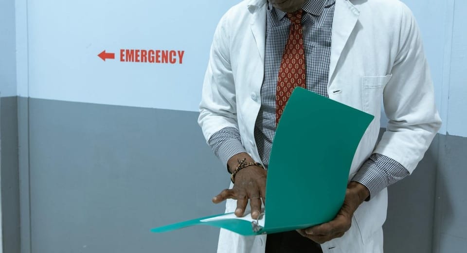 A doctor in a lab coat examines documents near the emergency room sign.
